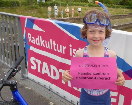 Ein Mädchen steht im Badeoutfit und Taucherbrille neben seinem Fahrrad. Im Hintergrund sind Schwimmer-Figuren zu sehen.