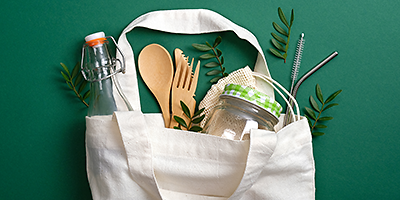 Eine wiederverwendbare Stofftasche liegt auf grünem Hintergrund. Darin enthalten ist Besteck aus Bambus, ein wiederverwendbarer Trinkhalt aus Metall sowie Glasbehälter.