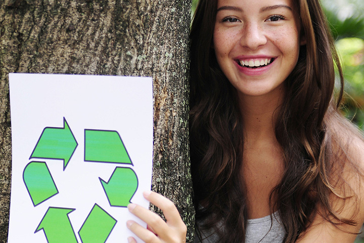 Junge Frau umarmt mit dem rechten Arm einen Baum und hält in der linken Hand ein Blatt mit Recycling-Logo