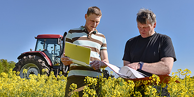 Zwei Männer stehen in einem Rapsfeld, halten einen Aktenordner und ein Notebook in den Händen und unterhalten sich. Im Hintergrund steht ein Traktor.