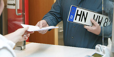 Ein Mann hält ein Kennzeichen und bekommt Fahrzeugpapiere überreicht