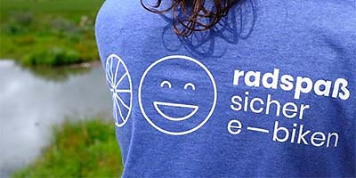 Eine Person trägt ein blaues T-Shirt mit weißer Aufschrift. Der Text lautet "radspaß sicher e-biken".